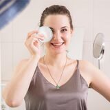Eine Frau steht im Badezimmer. Sie lächelt in die Kamera und hält sich dabei ein wiederverwendbares Wattepad vor ein Auge.