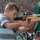 Ein Junge steht an einem Gewehr und zielt, dahinter ein Schütze in Uniform.