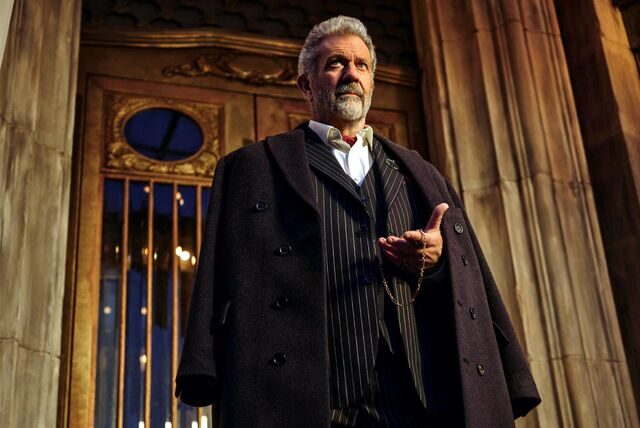 Mel Gibson in der Rolle des Hotel-Chefs Cornac O’Connor steht vor dem Eingang und blickt auf seine Taschenuhr.