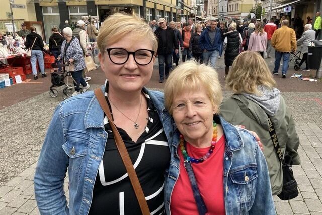 Zwei Frauen stehen nebeneinander auf der Hohe Straße und lächeln in die Kamera.Menschen flanieren zwischen Ständen auf der Hohe Straße in Datteln. Über ihren Köpfen ist ein Banner gespannt mit der Aufschrift „Welkom Holland-Markt“.