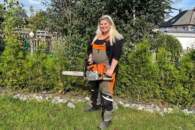 Eine Frau in Handwerkskleidung steht vor einer Hecke. In der Hand hält sie eine Kettensäge.