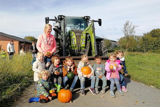 Kinder sitzen in der Schaufel eines Baggers, einige haben einen Kürbis in der Hand. Im Hintergrund steht eine Frau und lächelt.