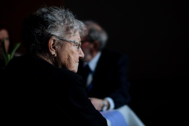 Ein Foto von zwei Personen, die an einem Tisch in einem schwach beleuchteten Raum sitzen. Die ältere Frau auf der linken Seite hat lockiges graues Haar und trägt eine schwarze Jacke. Sie trägt eine Brille, hat einen ernsten Gesichtsausdruck und schaut nach rechts. Die Person auf der rechten Seite trägt ein weißes Hemd und eine schwarze Jacke. Der Hintergrund ist dunkel und unscharf.