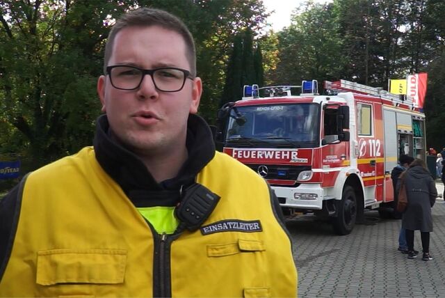 Ein junger Mann mit Feuerwehr-Montur steht vor einem Feuerwehr-Fahrzeug und beantwortet Fragen eines Reporters.