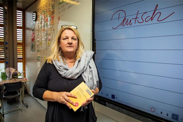 Stephanie Brzoza steht vor einer digitalen Tafel. Auf der Tafel steht in Schreibschrift „Deutsch“. In der Hand hält sie einen Duden.