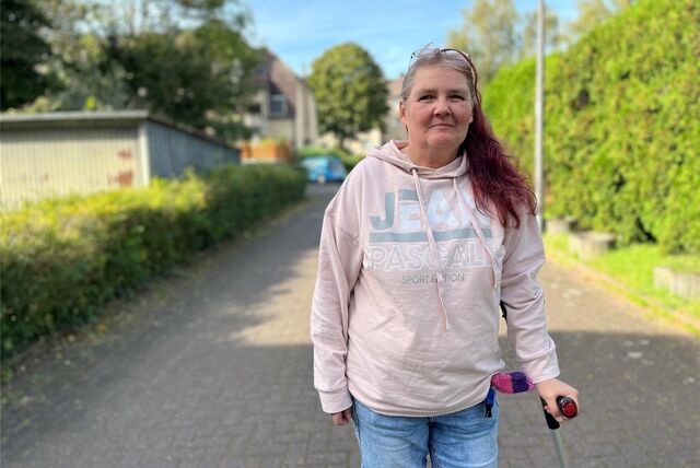 Das Bild zeigt eine Frau, die auf einer Straße geht. Die Person trägt einen hellrosa Kapuzenpullover mit der Aufschrift “JEANS” und blaue Jeans. Die Person hat eine Krücke. Der Hintergrund besteht aus einer Straße mit Häusern und Bäumen auf beiden Seiten.