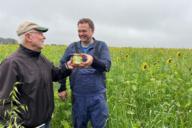 Udo Pfingst, links, übergibt Heiner Wessel ein Honigglas. Beide stehen in einem Sonnenblumenfeld.