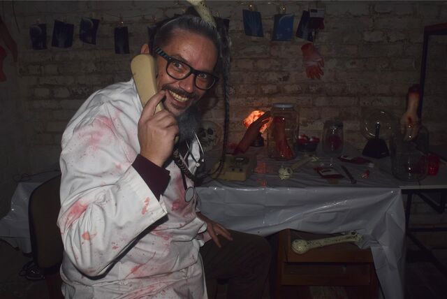 Ein Mann in einem weißen Kittel, der mit Kunstblut verschmiert ist, hält einen Telefonhörer in der Hand.