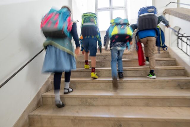 Schüler laufen eine Treppe hoch, man sieht sie von hinten, alle haben einen Schulranzen auf dem Rücken.