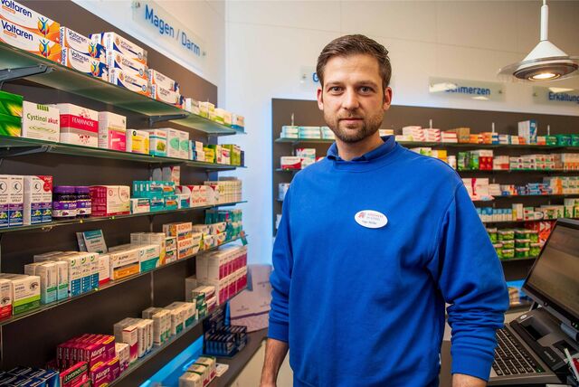 Ingo Müller steht vor Regalen, die mit Medikamentenpackungen gefüllt sind. Er trägt einen blauen Pulli mit einem Namensschild und dem Apothekensymbol.