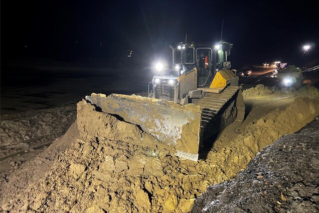 Ein beleuchtetes Baustellenfahrzeug schaufelt in der Nacht auf einer Baustelle Schlamm.