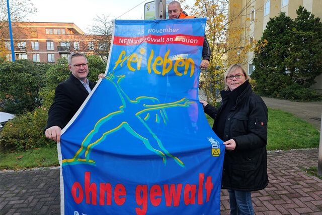 Drei Menschen halten eine Fahne mit der Aufschrift „frei leben ohne Gewalt hoch“.