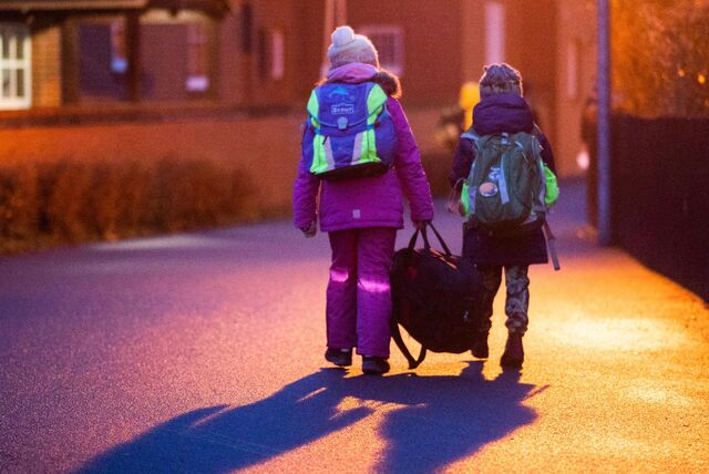 Das Bild zeigt zwei Kinder, die morgens eine Straße entlang gehen. Sie tragen Rucksäcke und tragen Taschen. Die Straße wird von Straßenlaternen beleuchtet, die ein orangefarbenes Leuchten erzeugen. Im Hintergrund sieht man ein Haus und einen Zaun. Die Kinder gehen auf dem Gehweg, der von einem kürzlichen Regen nass ist.