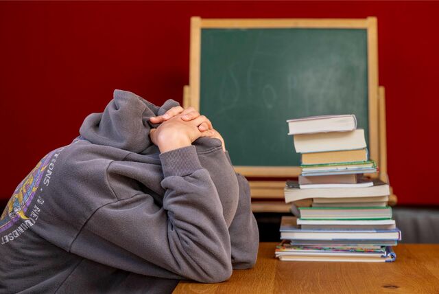 Eine Schülerin sitzt vor einem Stapel Bücher und hat die Kapuze ihres Pullovers über den Kopf gezogen.