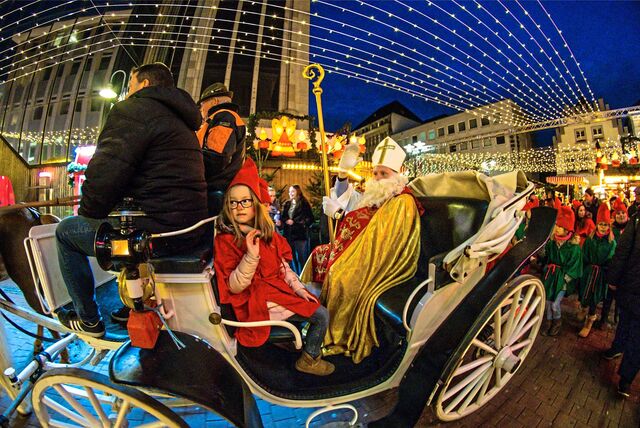 Ein als Nikolaus verkleideter Mann und ein Mädchen mit Wichtelmütze sitzen in einer weißen Kutsche. Sie winken und fahren über den Recklinghäuser Altstadtmarkt.