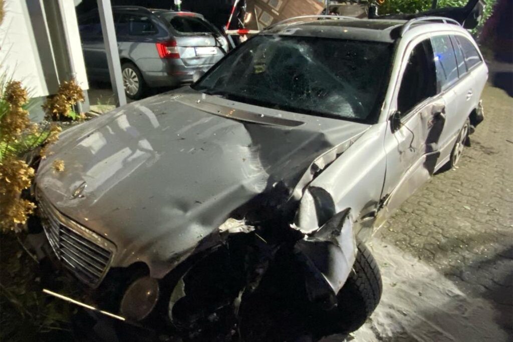 Ein graues Unfallauto steht in einem Vorgarten. Dahinter ein Fahrzeug in einem Carport. Ein graues Unfallauto steht in einem Vorgarten. Dahinter ein Fahrzeug in einem Carport.