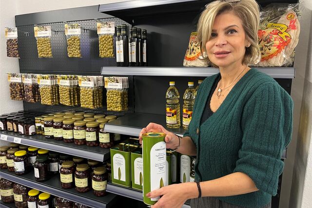 Sofia Chaita steht vor einem Supermarktregal mit diversen Gläsern voller Oliven und Flaschen mit Öl. In ihren Händen hält sie eine große Dose mit Olivenöl.