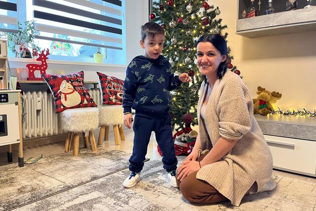 Noel Goxhaj und seine Mutter Esliona Cangollari (32) posieren vor dem Weihnachtsbaum im Wohnzimmer für ein Foto. Der Dreijährige steht selbstständig, während seine Mutter neben ihm kniet.