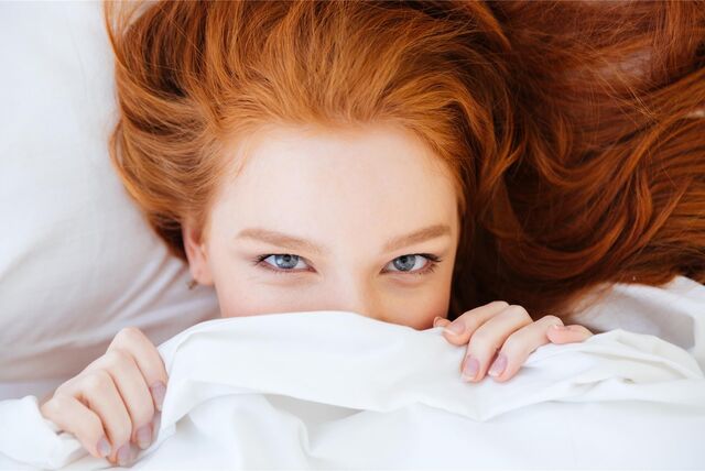 Rothaarige Frau versteckt sich unter der Bettdecke.