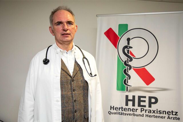 Klaus Hillebrand steht in weißem Arztkittel und Stetoskop vor einem Aufsteller des Hertener Praxisnetzes. Das Logo nimmt fast die gesamte rechte Hälfte des Bildes ein.