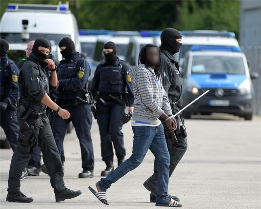 In der Landeserstaufnahmeeinrichtung für Flüchtlinge (LEA) wird ein gefesselter Mann von maskierten Polizisten abgeführt.