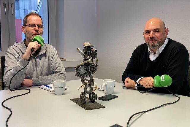 Zwei Männer haben jeweils ein Mikrofon in der Hand, sitzen an einem Tisch und lächeln in die Kamera.