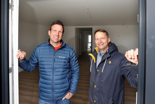 Zwei Männer in blauen Winterjacken und mit kurzen Haaren stehen an einer geöffnet Balkontür im Inneren einer Wohnung. Sie lächeln in die Kamera.