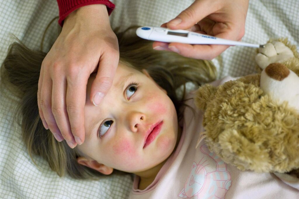 Ein kleines Mädchen liegt mit geröteten Wangen im Bett. Eine Mutter legt die Hand auf den Kopf und schaut auf ein Fieberthermometer. Ein kleines Mädchen liegt mit geröteten Wangen im Bett. Eine Mutter legt die Hand auf den Kopf und schaut auf ein Fieberthermometer.