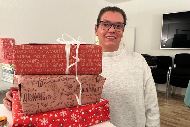 Erzieherin Yvonne Schweda von der Kita Kinderplanet lächelt. Die trägt einen weißen Pullover und eine schwarze Brille. Vor ihr auf dem Tisch liegen drei Weihnachtsgeschenke, verpackt in rotem Papier.