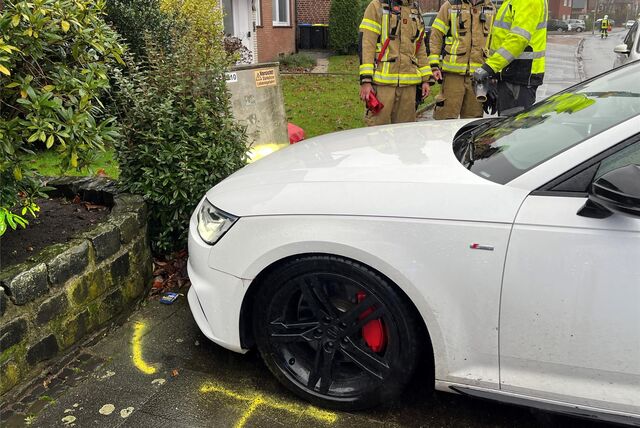 Ein weißes Auto steht auf einem Bordstein. Im Hintergrund stehen Feuerwehrleute.