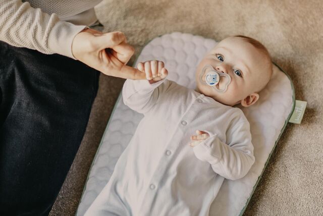 ein Baby, das auf einer weichen Unterlage liegt und einen Schnuller im Mund hat. Eine Person, von der nur ein Teil zu sehen ist, hält die Hand des Babys. Das Baby trägt einen weißen Strampler und schaut direkt in die Kamera. Der Hintergrund ist unscharf und zeigt eine weiche, texturierte Oberfläche.