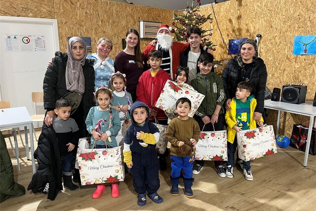 Eine Gruppe von Menschen steht zusammen mit dem Weihnachtsmann und Eiskönigin Elsa vor einem Weihnachtsbaum und posiert für ein Erinnerungsfoto. Viele der Kinder haben große und mit Geschenken befüllte Weihnachtstüten in der Hand.
