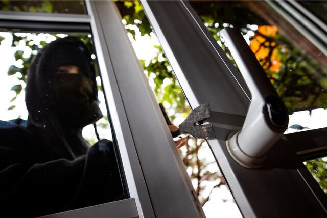 Schwarz vermummter Einbrecher steckt sein Brecheisen zwischen zwei Fensterflügel und wird von innen fotografiert. Der Fenstergriff ist nach oben gerichtet.