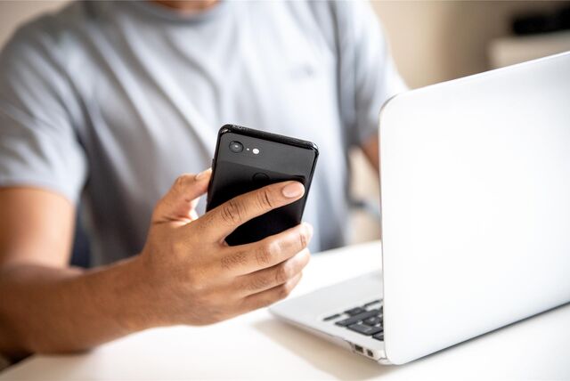Ein Mann sitzt an seinem Laptop und hält sein Handy in der Hand.