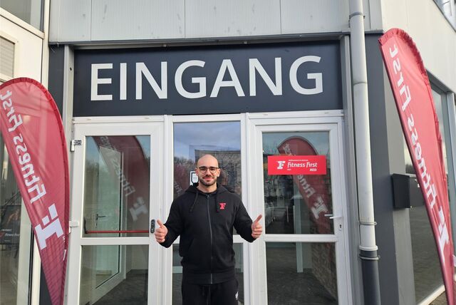 Ein Fitnessstudio-Mitarbeiter steht vor dem Gebäude. Dort steht Eingang.