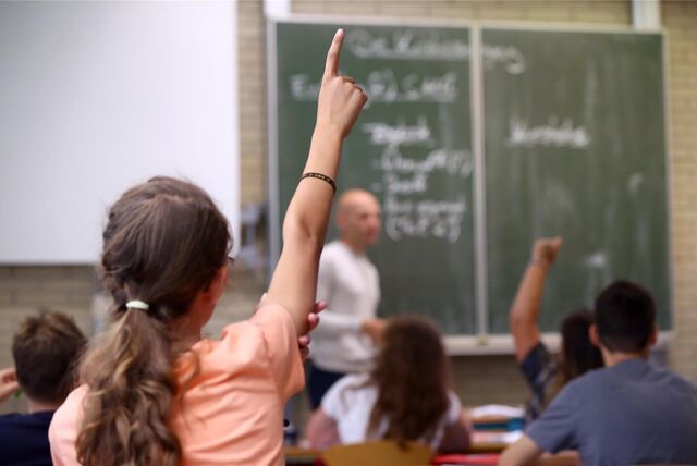 Blick in eine Klasse: Ein Mädchen zeigt auf, an der Tafel steht ein Lehrer.