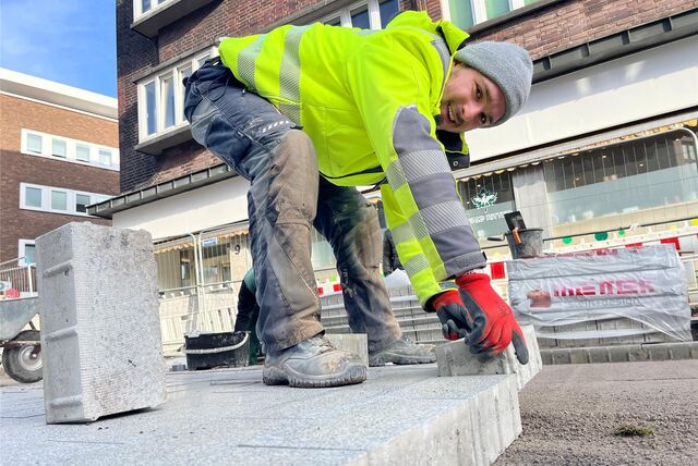 Froschperspektive: Bauarbeiter in gelber Warnweste steht auf der bereits neue gepflasterten Fläche, beugt sich tief nach unten und legt mit roten Schutzhandschuhen einen Pflasterstein an die anderen.