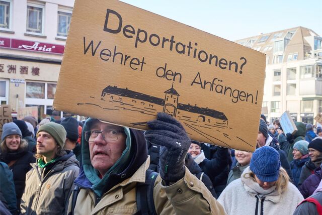 Menschen protestieren gegen Rechts. Ein Mann hält ein Schild mit einer Zeichnung des Konzentrationslagers Auschwitz in die Höhe. Darüber die Worte „Deportationen? Wehret den Anfängen!“