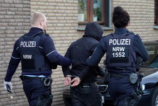 Ein Mann mit Kapuze auf dem Kopf wird von zwei Polizisten abgeführt.