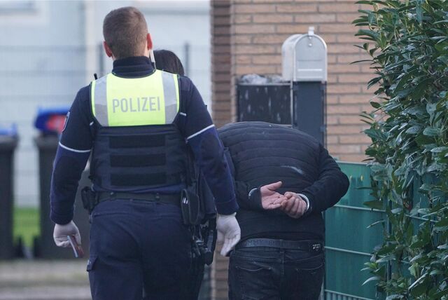 Ein Polizist führt einen Verhafteten ab. Der Mann hat die Hände hinter seinem Rücken in Handschellen und läuft gebückt.