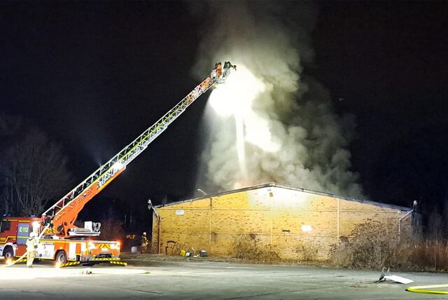 Eine Drehleiter der Feuerwehr steht vor einer Lagerhalle. Rauch steigt aus dem Gebäude auf.
