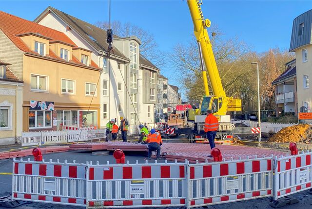 Ein gelber Kran hebt ein Teilstück eines Kreisverkehrs ein. Ein paar Bauarbeiter richten das Teilstück aus.