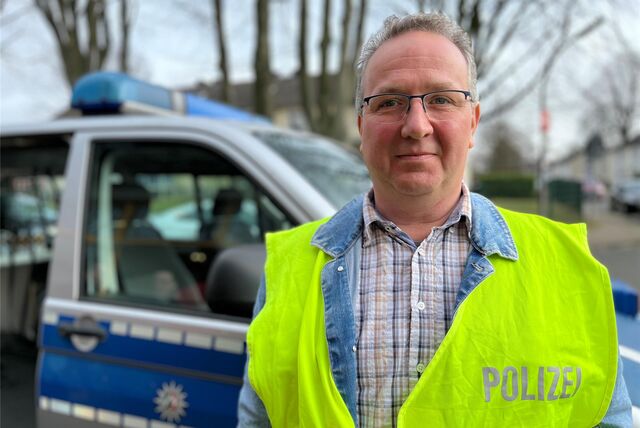 Polizist Andreas Schink trägt eine Warnweste. Er steht vor einem Polizeiwagen.