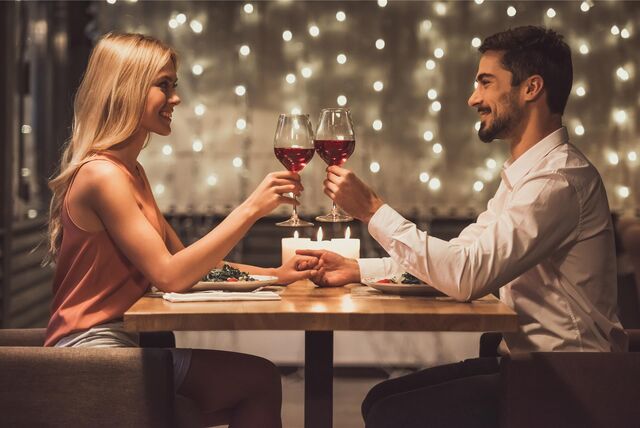 Eine blonde Frau sitzt an einem kleinen Tisch gegenüber von einem jungen Mann. Sie heben ihre Weingläser, um anzustoßen.