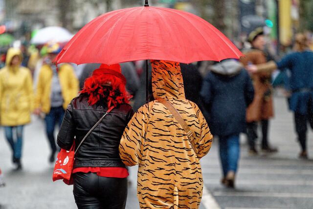 Unter einem großen roten Regenschirm laufen zwei Karnevalisten. Eine Frau trägt Lederkleidung mit roten Akzenten und einer Federboa. Ihre Begleitung hat einen Tiger-Einteiler an.