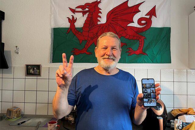 Detlef Kihm steht in seiner Küche vor der walisischen Flagge und hält sein Handy in die Kamera.