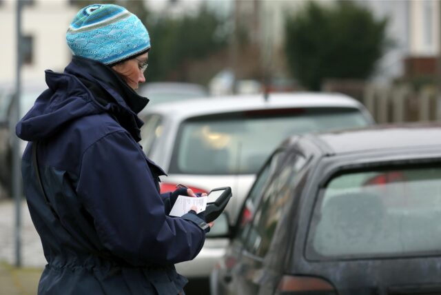 Eine Politesse steht neben mehreren geparkten Autos und stellt einen Strafzettel aus.