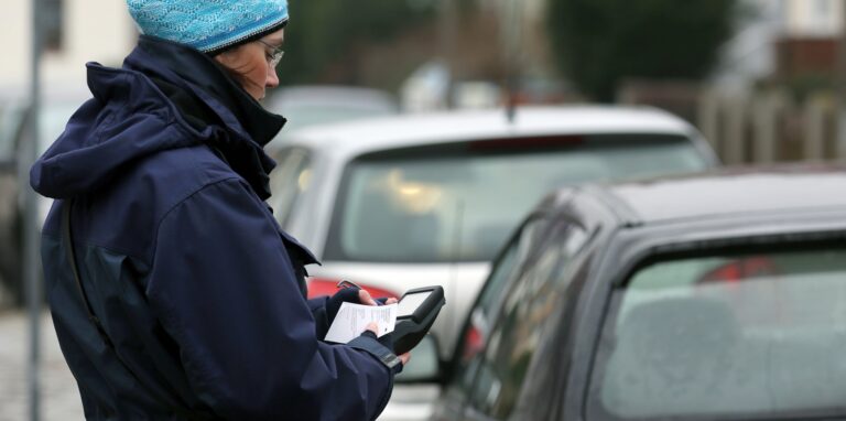 Eine Politesse steht neben mehreren geparkten Autos und stellt einen Strafzettel aus.