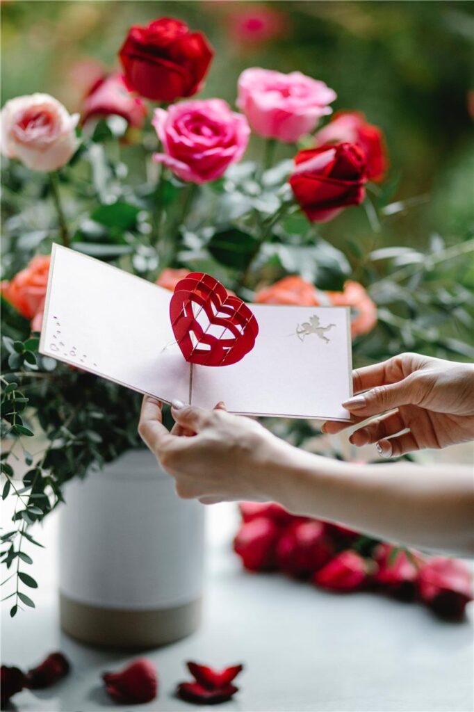 Eine Frau hält eine Karte zum Valentinstag in den Händen, dahinter steht ein Blumenstrauß. Eine Frau hält eine Karte zum Valentinstag in den Händen, dahinter steht ein Blumenstrauß.