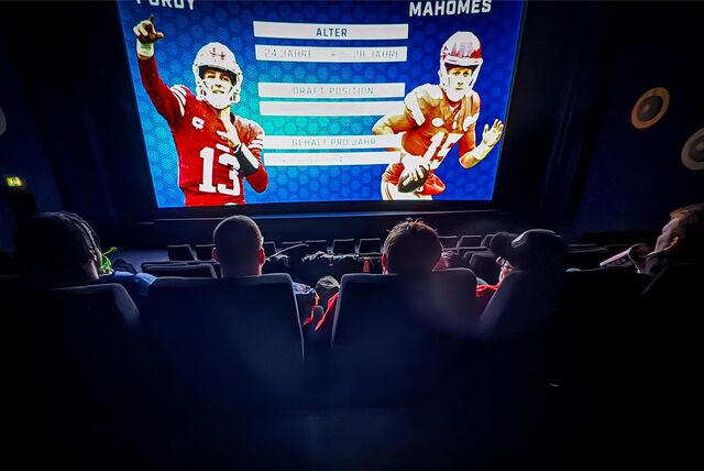 Zuschauer sitzen in Kinosesseln. Auf der Leinwand sind zwei Footballspieler zu sehen.
